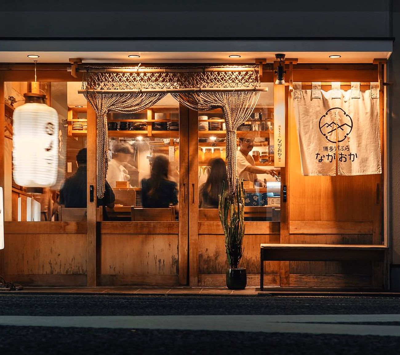 通りから見た夜の博多天ぷら ながおか本店の外観。提灯とのれん、店内で食事するお客様